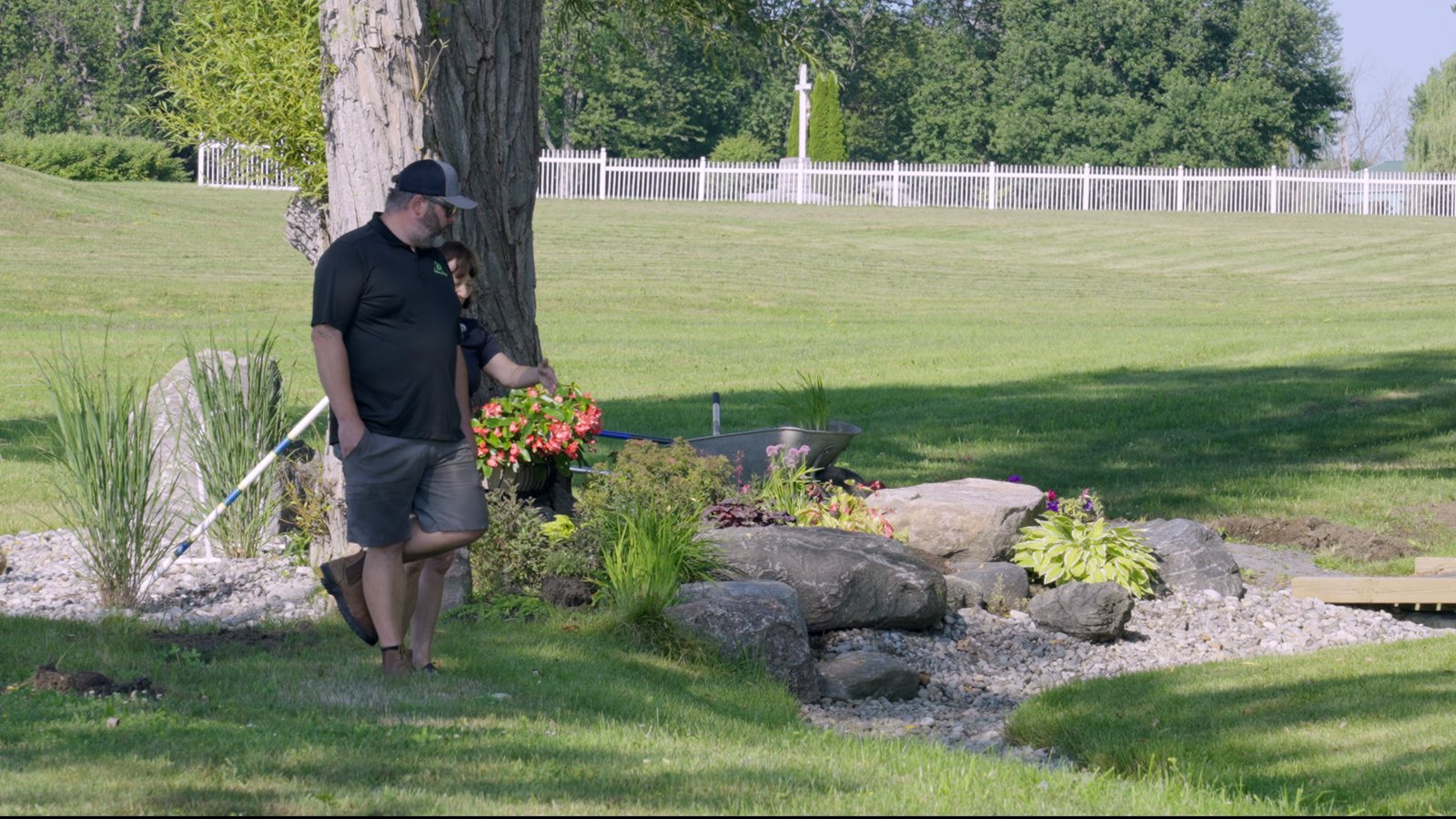 A man tending a planted bed set with stone beside a lawn and mature trees